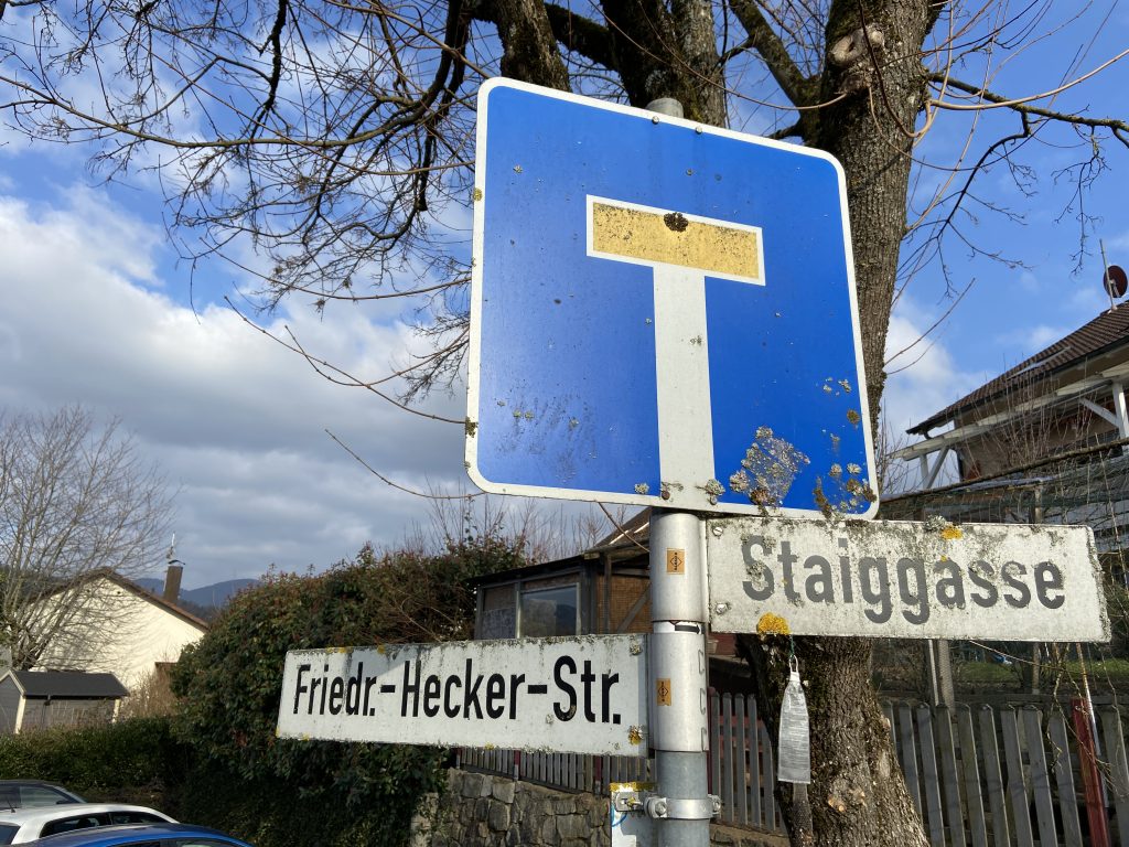 Schild einer Sackgasse mit blauem Verkehrszeichen „Ende der Straße“ über Straßenschildern für „Friedr.-Hecker-Str.“ und „Staiggasse“, vor einem Baum, Häusern und bewölktem Himmel.