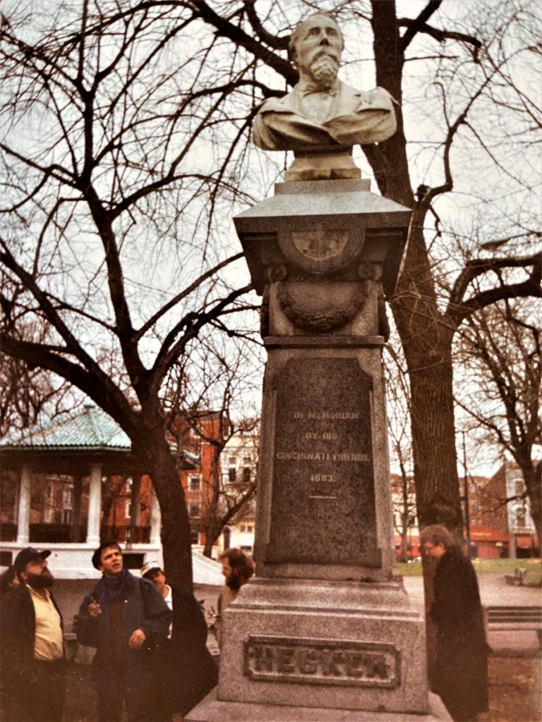 Fotografie des Friedrich-Hecker-Denkmals in Washington Park, Cincinnati: Eine steinerne Büste von Friedrich Hecker auf hohem Sockel, umgeben von Parkbäumen und einigen wenigen interessiert wirkenden Besucher*innen.