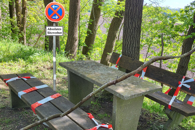 Das Foto zeigt zwei Holzbänke und einen steinernen Tisch im Wald, um die Sperrband gewickelt ist. Im Hintergrund ist ein Schild mit der Aufschrift „Absolutes Sitzverbot“.
